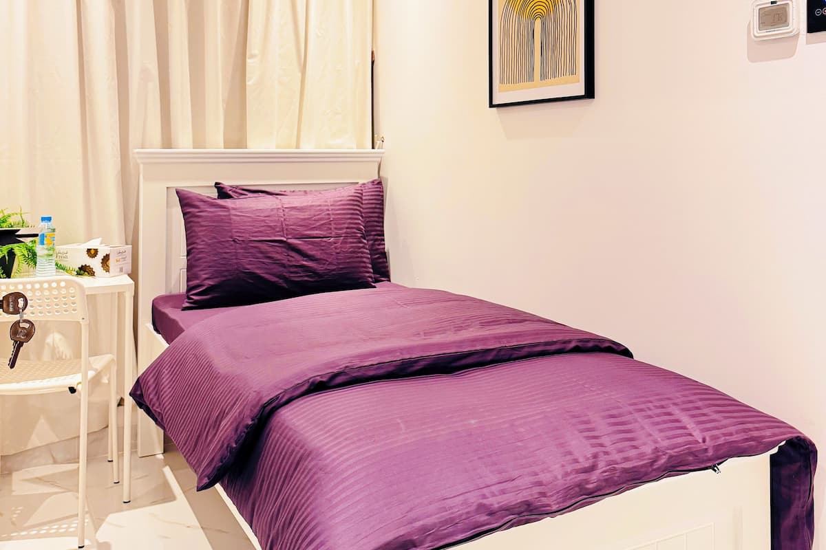 A bedroom with a bed, a nightstand, a chair, a window with curtains, a wall-mounted air conditioner, a framed picture, and a thermostat.