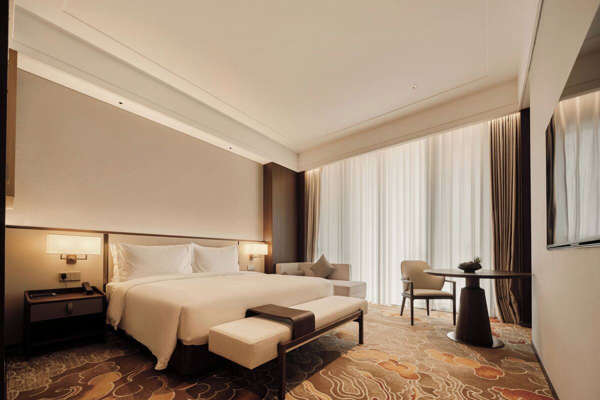 Habitación de hotel con una cama grande, una mesita de noche, una silla, una pequeña mesa y un amplio ventanal con cortinas.