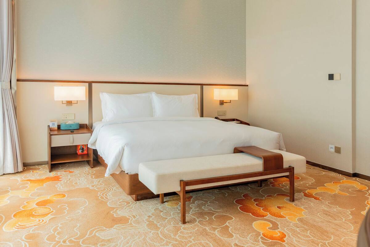 Una habitación de hotel con cama, lámparas de noche, mesita de noche, un banco y una alfombra estampada.