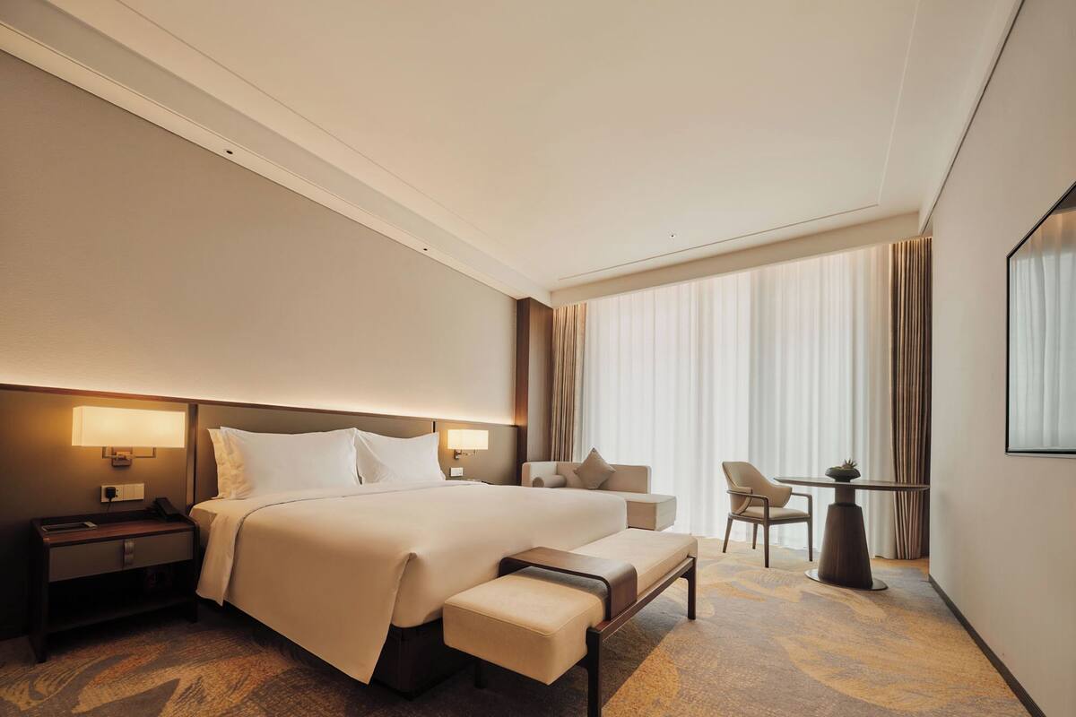 Habitación de hotel con una cama grande, una mesita de noche, una silla, una pequeña mesa con una planta y un ventanal con cortinas.