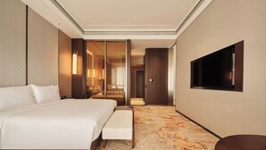 Une chambre d’hôtel moderne avec un grand lit, un banc, un téléviseur fixé au mur et un couloir recouvert de moquette.