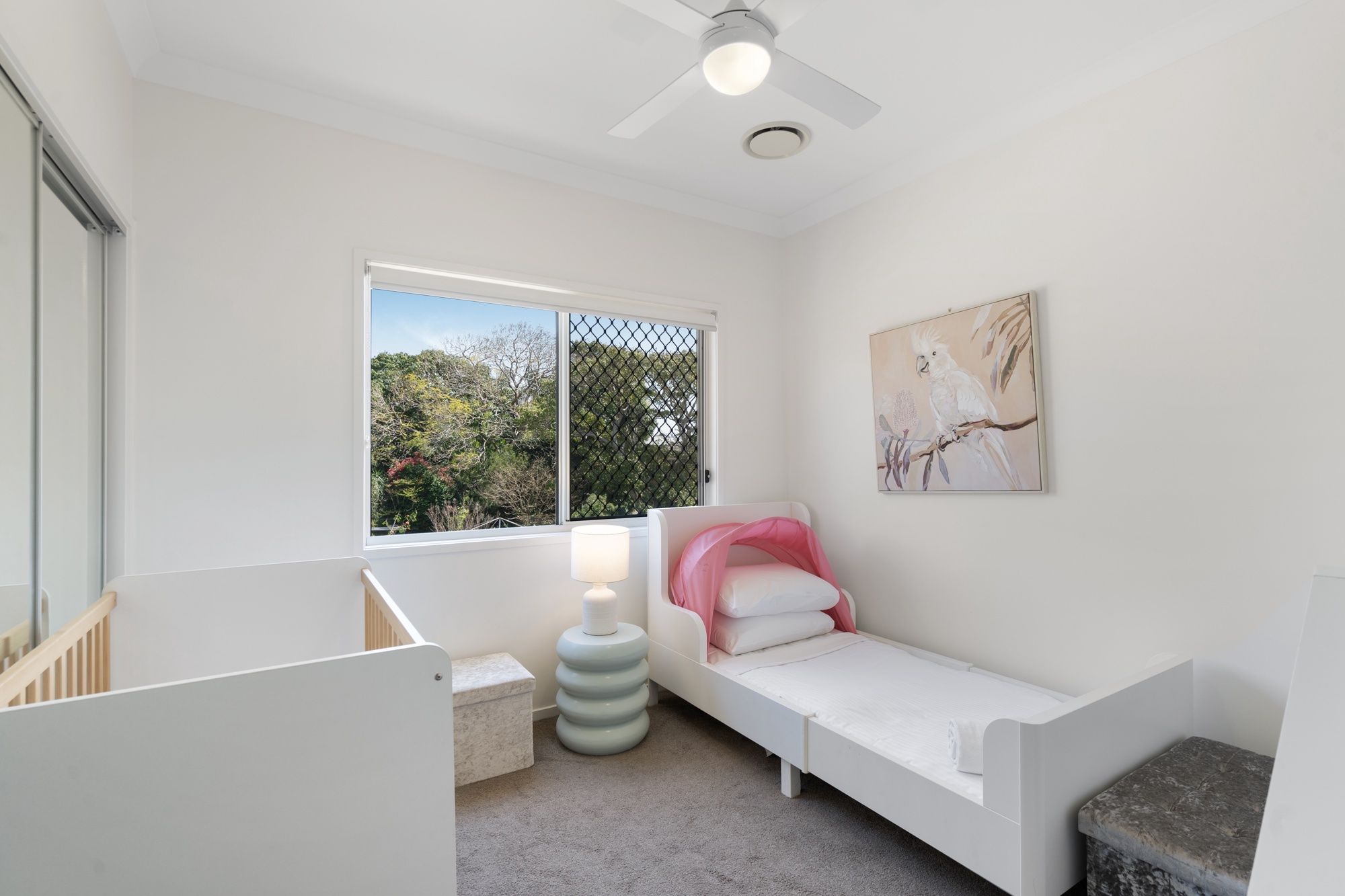 Un dormitorio con una cama individual, una ventana con vista a árboles, un ventilador de techo y un cuadro en la pared.