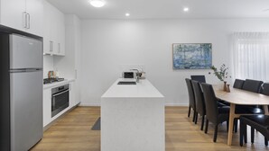 A modern kitchen with white cabinetry, a central island, and a dining area with a table and chairs. - Family-Friendly House with Courtyard Retreat (Wavell Heights)
