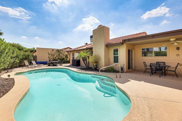 A swimming pool with a curved edge, a patio area with a table and chairs, and a covered outdoor seating area. - Golf, Swim & Grill: Snowbird’s Escape in Sun Lakes (Sun Lakes)