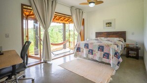 A bedroom with a bed, a desk, a chair, a hammock, and a view of greenery through the window.