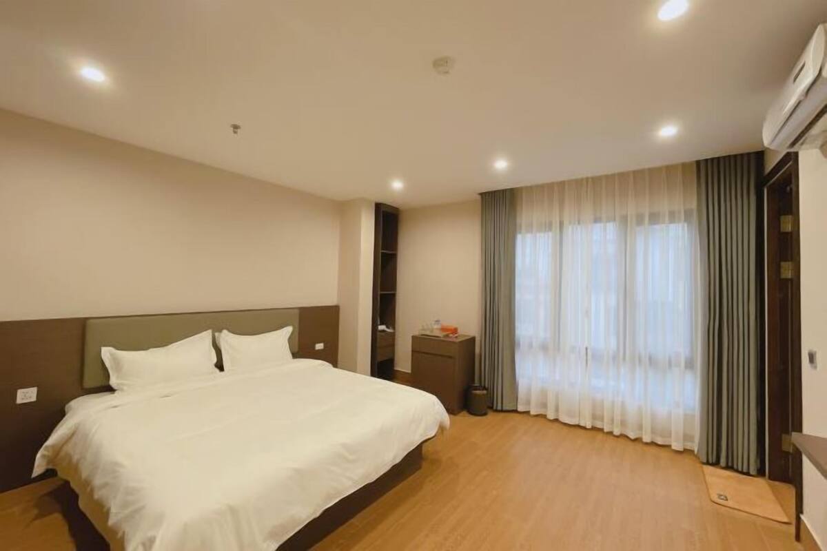 A hotel room with a bed, a nightstand, a window with curtains, and an air conditioning unit.