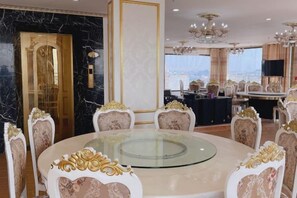 A luxurious dining area with a round glass table surrounded by ornate chairs, a chandelier, and a view of the city. - KIM A HOTEL (Bac Ninh)