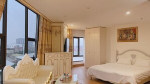 A spacious bedroom with a large bed, a sofa, and a wardrobe. There is a painting on the wall and a window providing a view of the city.