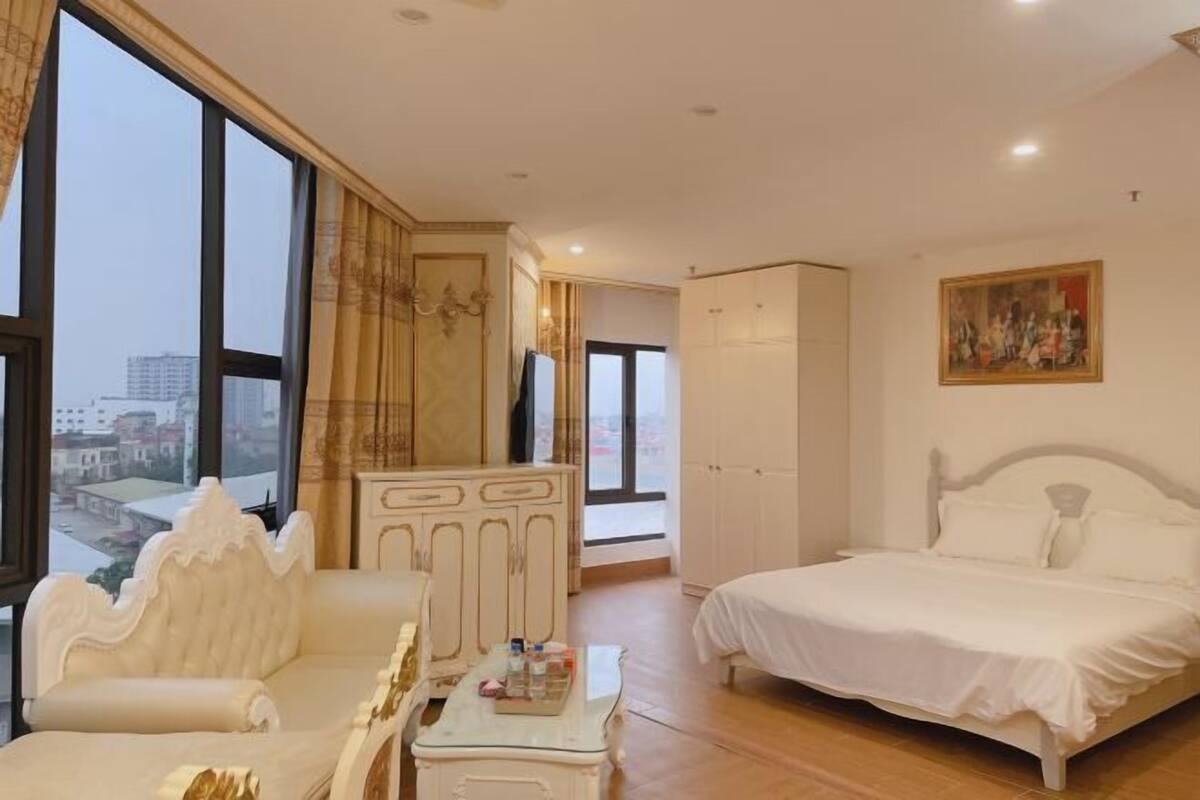 A spacious bedroom with a large bed, a sofa, and a wardrobe. There is a painting on the wall and a window providing a view of the city.