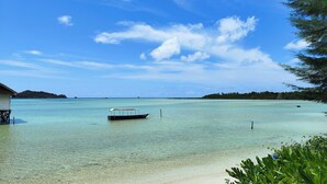 A clear tropical beach with a boat in the water, a small hut on stilts, and a forested island in the background. - Roka Resort by ARKasia Foundation (Bintan)