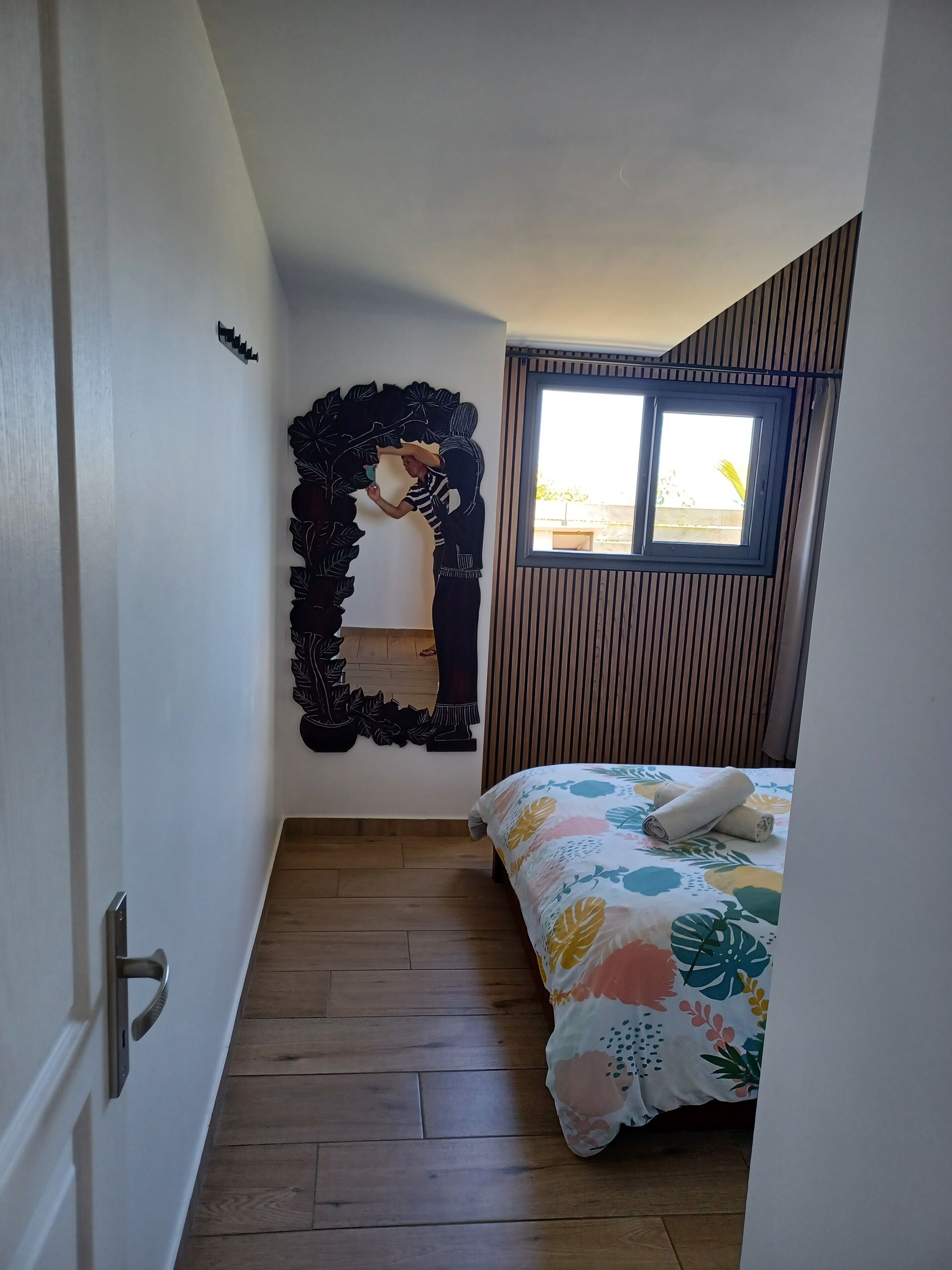 A bedroom with a bed, a window, a wooden floor, and a decorative mirror.