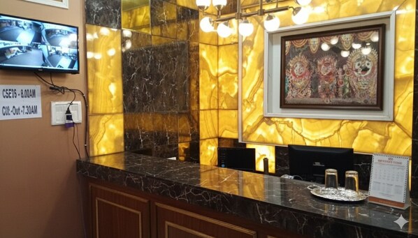 A hotel reception area with a marble counter, a television displaying security footage, a framed artwork, and a calendar on the wall. - Sahoo Palace (Brahmagiri)