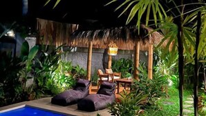 Un espace piscine agrémenté de chaises longues et d’une table, entouré d’une végétation luxuriante et d’une structure à toit de chaume.