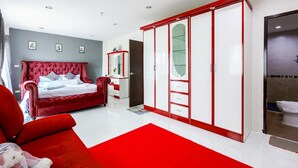 A bedroom with a red upholstered bed, a red chair, a wardrobe with a mirror, and a bathroom visible in the background.
