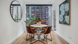 A modern dining area with a round table, four brown chairs, a vase with flowers, a large window with a city view, and a painting on the wall. - Spacious City Retreat with Study (Victoria)