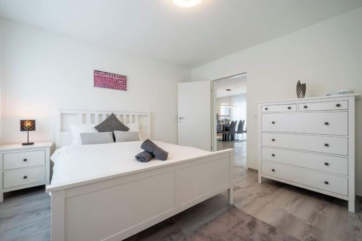 Un dormitorio con cama, mesitas de noche, cómoda y puerta que da a otra habitación.