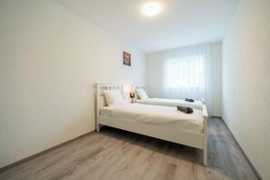 Un dormitorio con dos camas individuales, una mesita de noche, una ventana con cortinas translúcidas y suelo de madera.