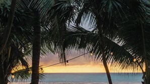 A beachside seating area with a cushioned bench, a small table, and a hammock under palm trees, overlooking the ocean at sunset. - Villa La Semilla • 5BR Beachfront w/ Chef (Tulum)