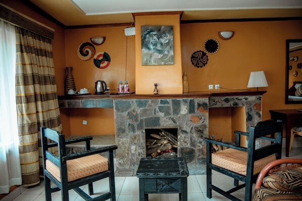 A cozy living room with a stone fireplace, two wooden chairs, a coffee table, and a wall decorated with various art pieces. - Mountain Gorilla View Lodge (Kinigi)