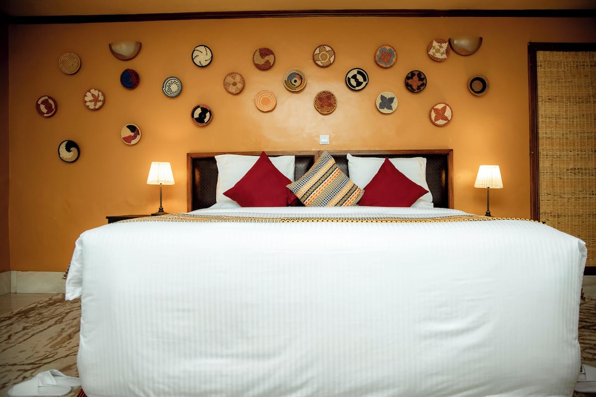 A hotel room with a large bed, two red pillows, a patterned throw, and a wall decorated with various woven plates.