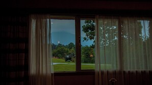 A room with a large window, a table with a vase, and a view of a grassy area with a statue. - Mountain Gorilla View Lodge (Kinigi)