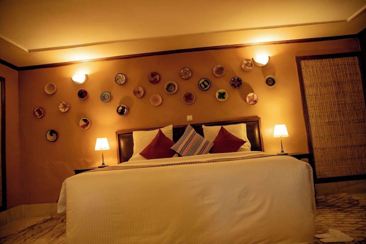 A hotel room with a bed, a headboard with decorative pillows, wall-mounted lights, and a large window with a view.