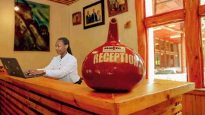 A reception desk with a large red vase labeled "RECEPTION" and a person using a laptop. - Mountain Gorilla View Lodge (Kinigi)