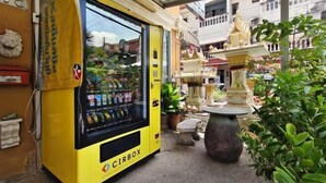 Ein gelber Verkaufsautomat mit der Aufschrift 'CIRBOX' im Freien, mit einer Statue und Blumentöpfen.