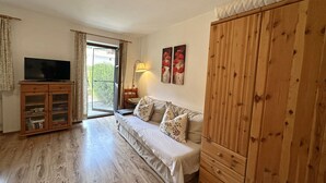A living room with a wooden floor, a sofa, a TV on a wooden cabinet, and a sliding glass door leading to a garden. - Apartment 38 sqm living/bedroom and terrace, App. 4 (Aschau im Chiemgau)