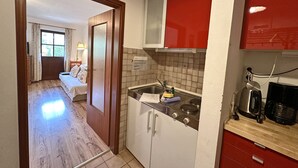 A compact hotel room with a wooden floor, a bed with white bedding, a small sitting area with a chair and lamp, and a kitchenette with a stove, sink, and red cabinets. - Apartment 38 sqm living/bedroom and terrace, App. 4 (Aschau im Chiemgau)