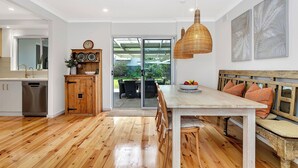 A modern kitchen with wooden flooring, a dining area with a bench and table, and a view through glass doors to an outdoor patio. - Daisy's Beachside Cottage-Picnic Garden, Pool Table & Games in Port Noarlunga (Port Noarlunga South)