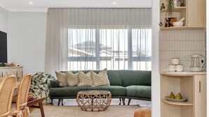 A living room with a green sofa, a round coffee table, and a dining area with wicker chairs. - Daisy's Beachside Cottage-Picnic Garden, Pool Table & Games in Port Noarlunga (Port Noarlunga South)