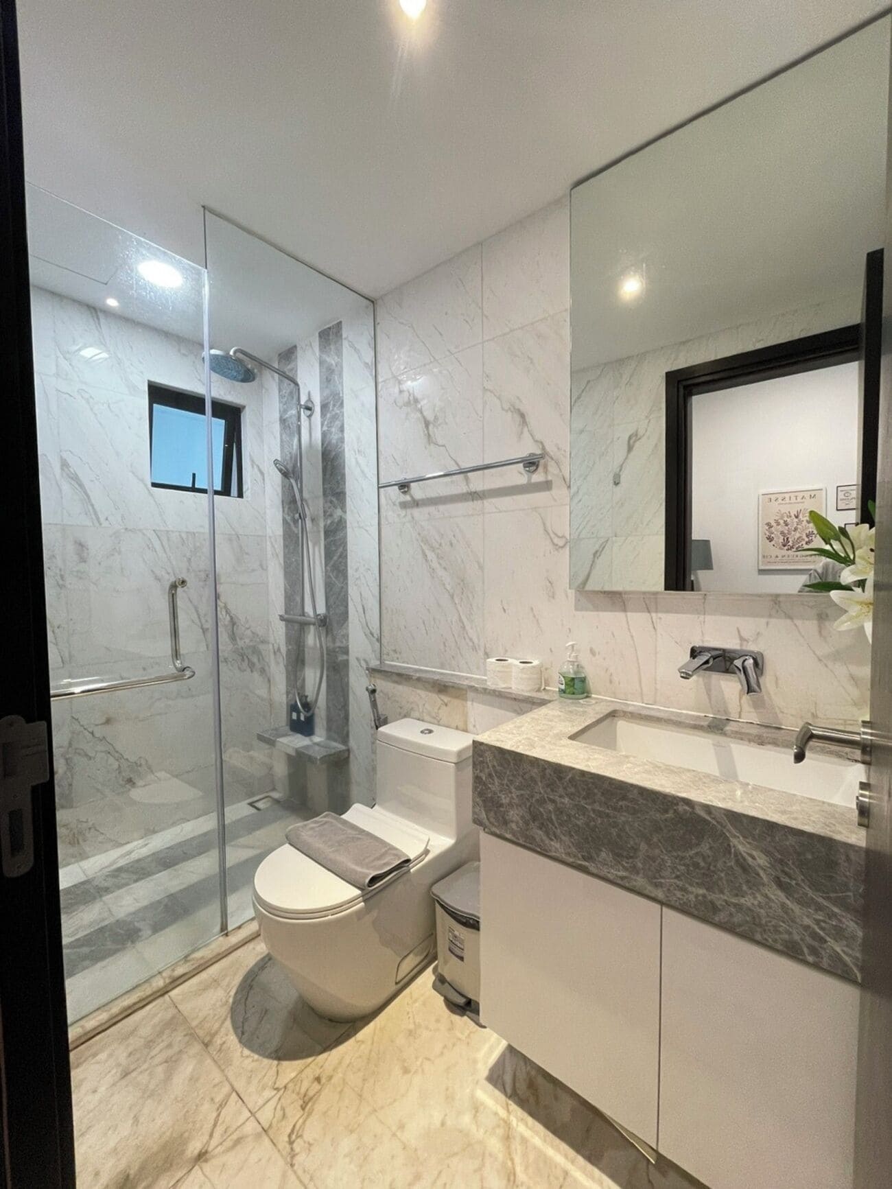 A modern bathroom with a glass-enclosed shower, a toilet, a sink with a marble countertop, and a large mirror.