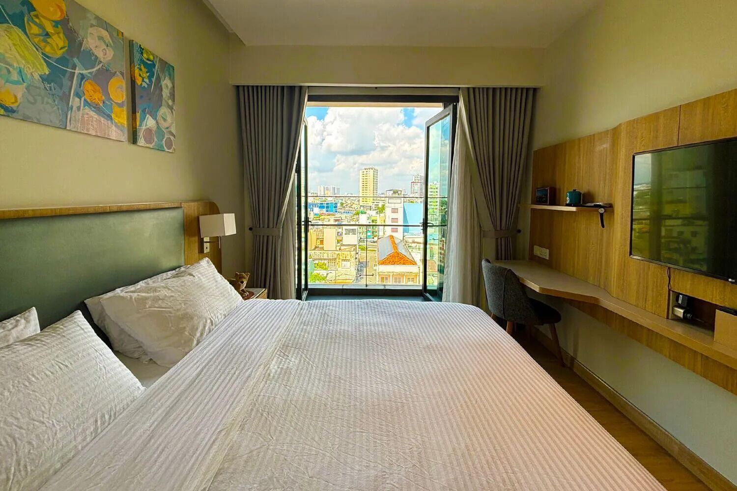 A hotel room with a large bed, a TV, a desk, and a view of the cityscape through the window.