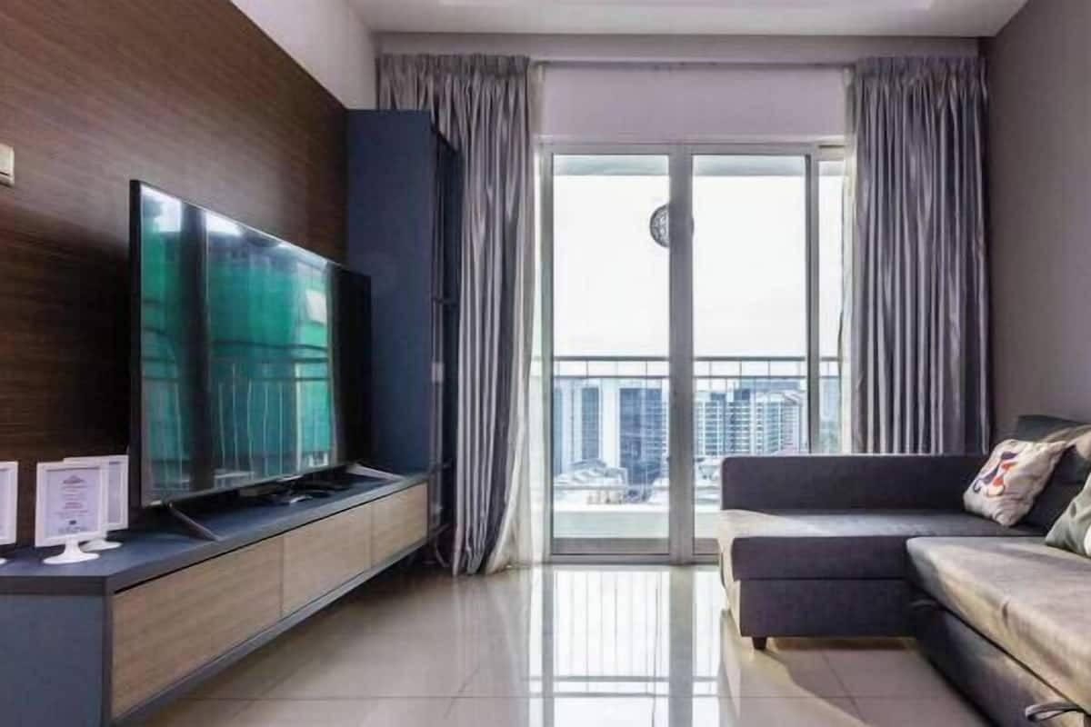 A modern living room with a flat-screen TV, a sofa, a sliding glass door leading to a balcony, and a city view.
