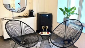 A modern room with two black wireframe chairs, a small table with two glasses of wine, a coffee machine, and a potted plant by the window.