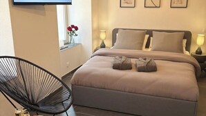 A bedroom with a bed, two bedside lamps, a TV on the wall, a chair, and a small table with a plate of food and drinks.