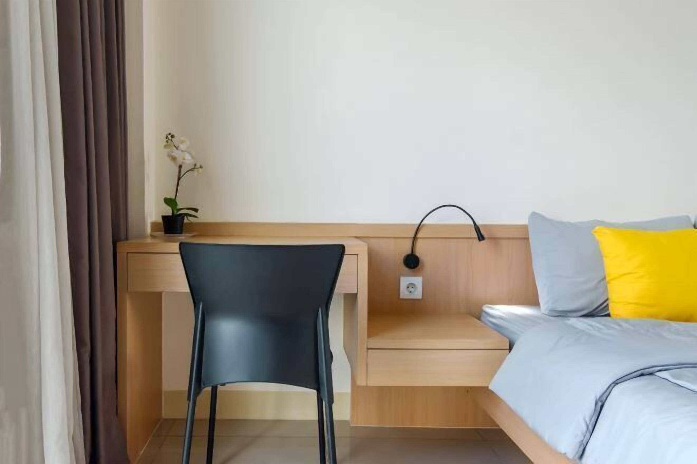 A hotel room with a wooden desk, a black chair, a small plant, a bedside table, and a bed with a grey and yellow pillow.