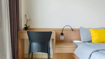 A hotel room with a wooden desk, a black chair, a small plant, a bedside table, and a bed with a grey and yellow pillow.