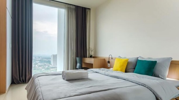 A hotel room with a large bed, a nightstand, a lamp, and a view of the city. - Gala by TwoSpaces Studio @TreePark Apartment (Serpong)