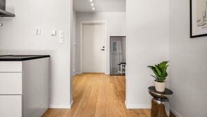 Una cocina moderna con armarios blancos y encimera negra, que da a un pasillo con suelo de madera, una puerta y una pequeña planta en maceta.
