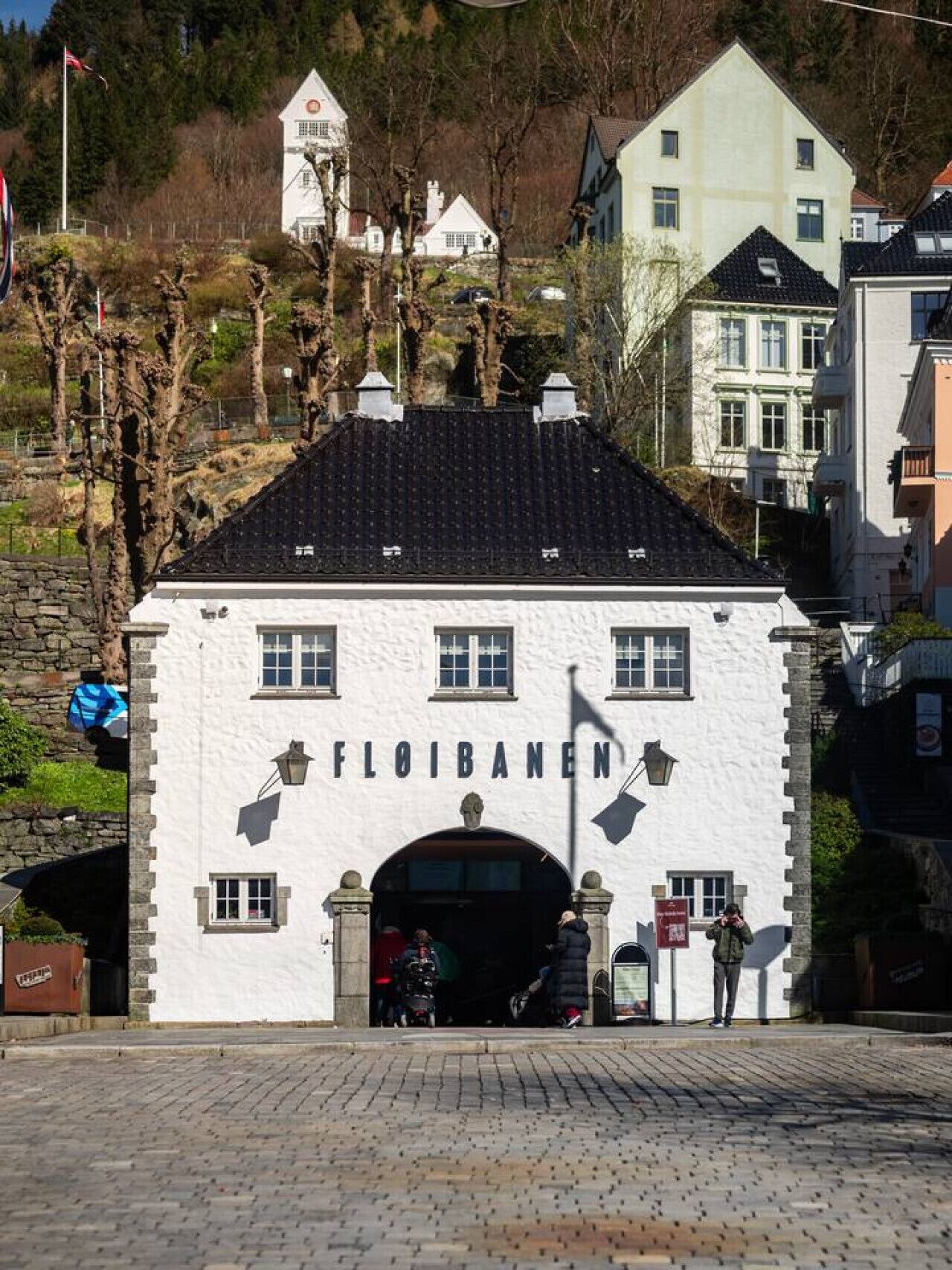 A white building with the name "FLOIBANEN" on the facade, featuring a stone entrance and a black tiled roof.