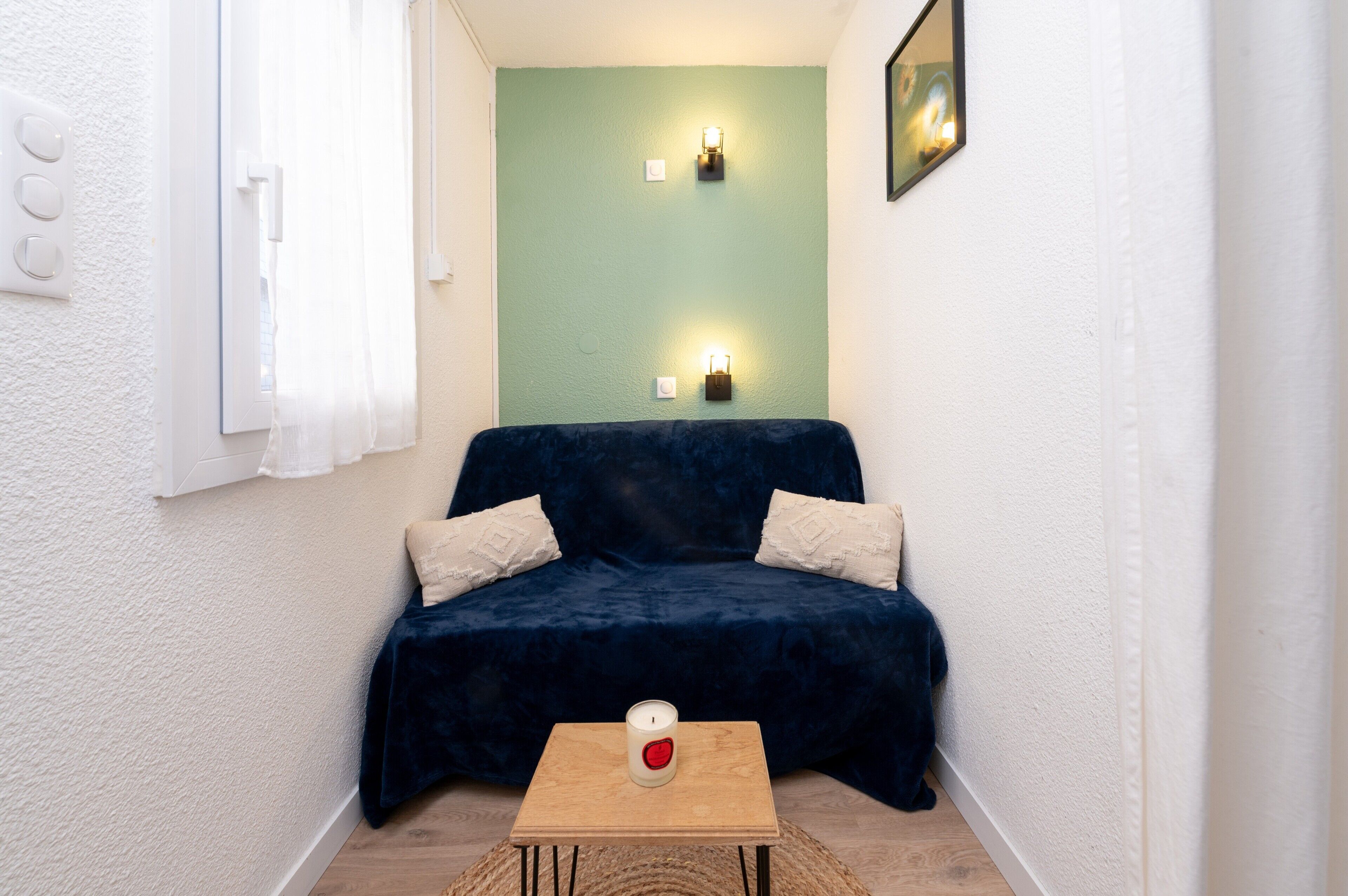A small room with a dark blue sofa, two white pillows, a wooden table with a candle, and a green accent wall.