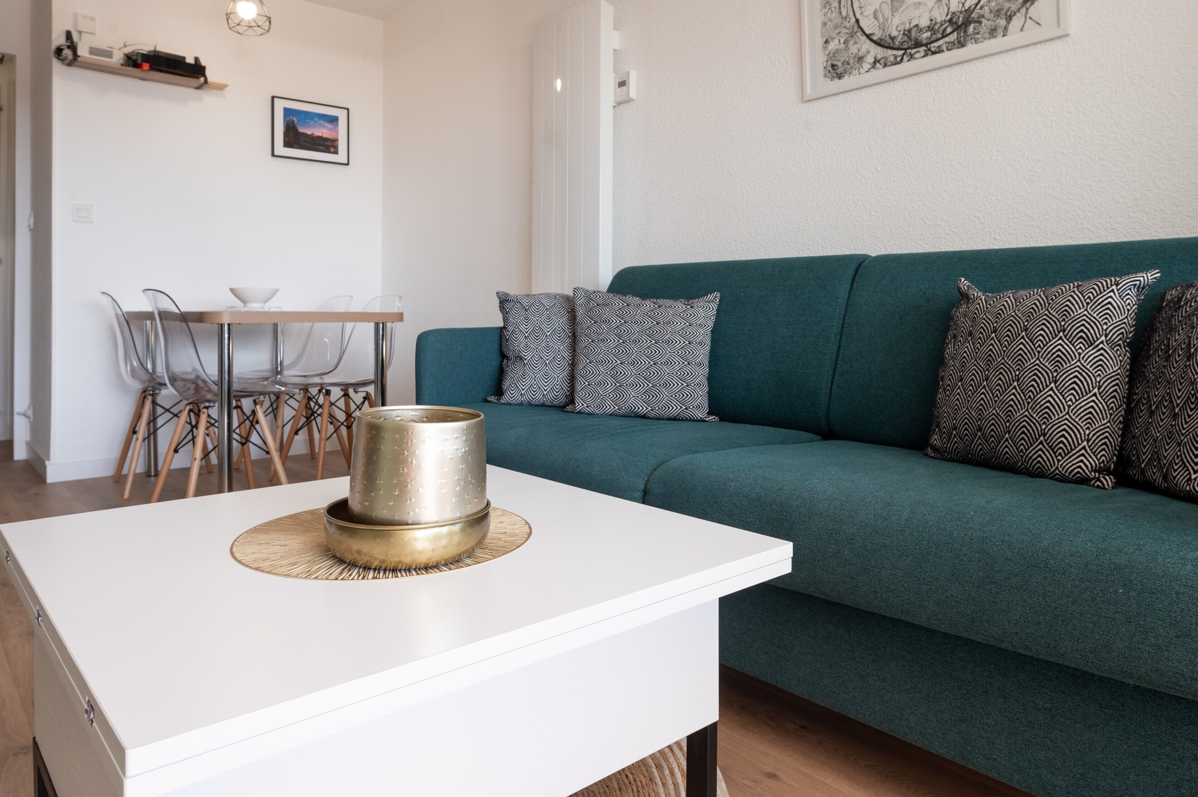 Un salon moderne avec une table basse blanche, un canapé vert agrémenté de coussins à motifs et un coin repas comprenant une table et des chaises en bois.