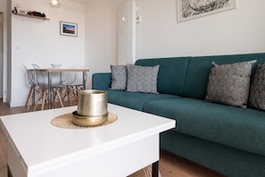 Un salon moderne avec une table basse blanche, un canapé vert agrémenté de coussins à motifs et un coin repas comprenant une table et des chaises en bois.