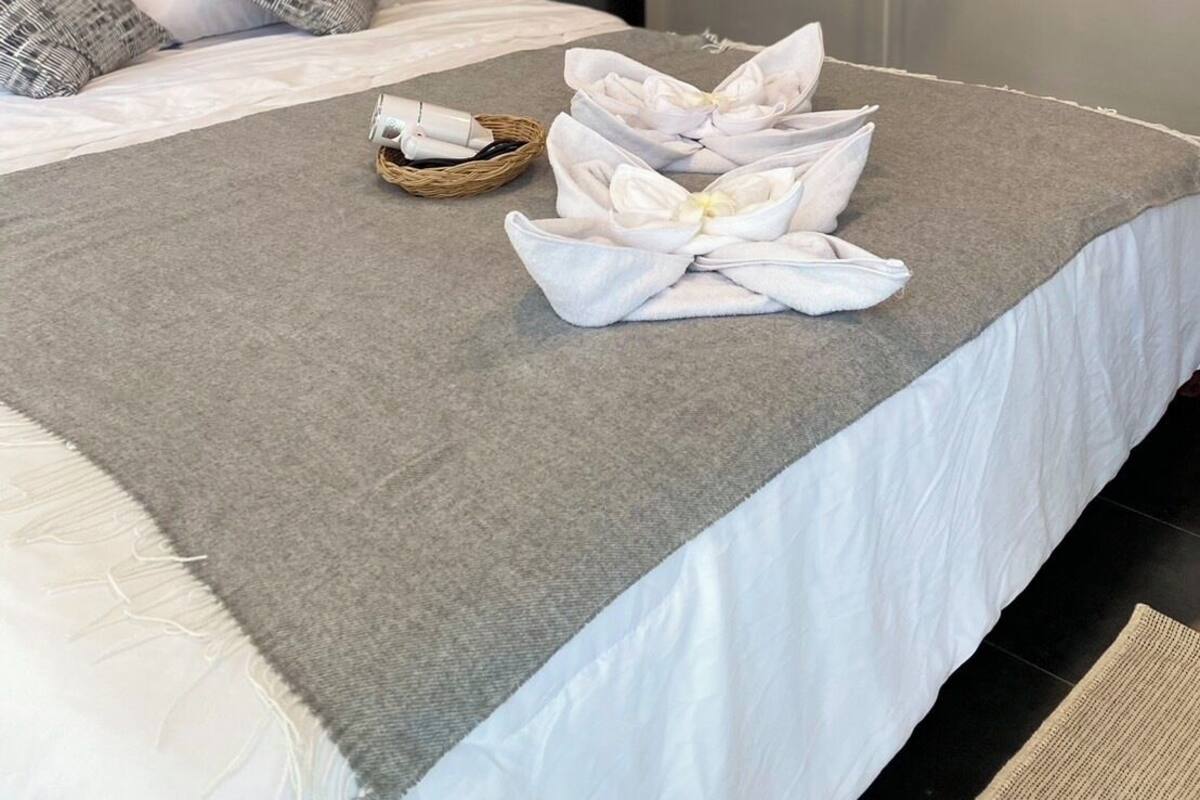 Una cama bien tendida con una manta gris, almohadas blancas y toallas dispuestas sobre ella.