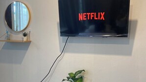 A wall-mounted television displaying 'NETFLIX' in red letters, a round mirror, a small shelf with a plant, and a bamboo stool. - Baaninkan (Ban Tai)