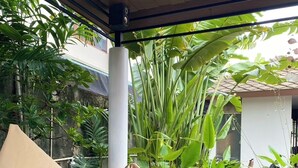 A beige bean bag chair on a wooden deck with a view of lush green plants and a building in the background. - Baaninkan (Ban Tai)