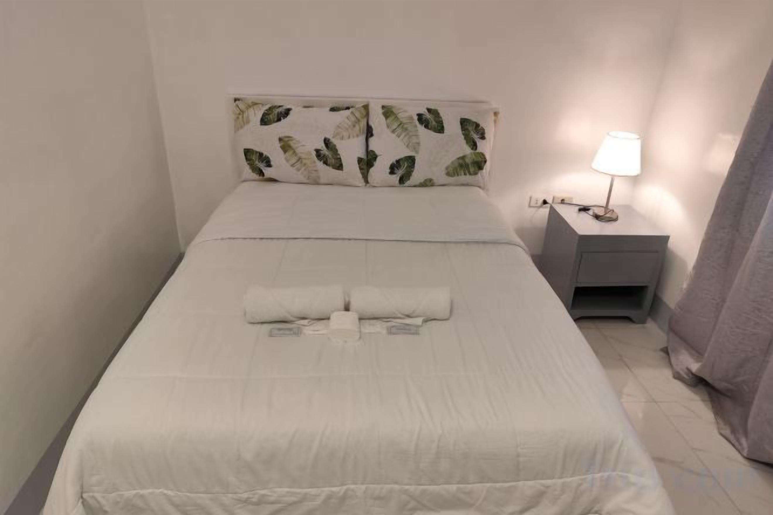A neatly made bed with white bedding and a pillow with a leaf pattern. There is a small bedside table with a lamp and a drawer.