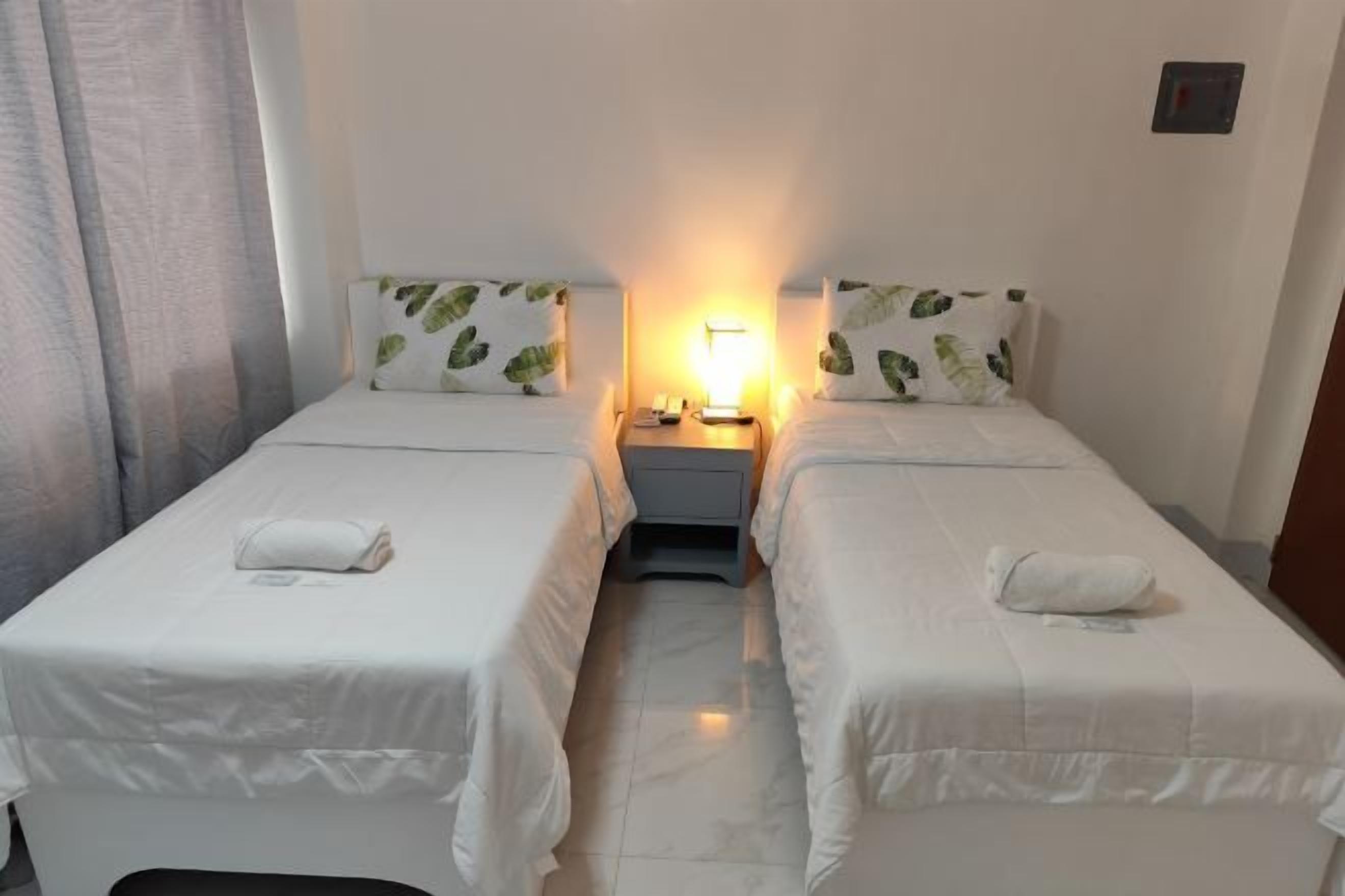A hotel room with two beds, a nightstand, a lamp, and a window with curtains.